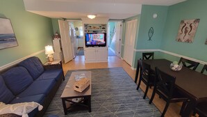 A living room with a blue sofa, a coffee table, a TV, and a dining area with a table and chairs. - Ocean Side Beach Condo in Family Friendly Community!  Oceanfront pool!! (Pine Knoll Shores)