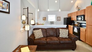 A cozy living room with a brown leather sofa, a wooden coffee table, and pendant lights. A kitchen with wooden cabinets and appliances is visible in the background. - Carriage House Cutie (KING BED) -Steps From Lake & Park (Bozeman)
