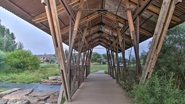 A covered wooden bridge with a metal railing, leading to a grassy area with a river and houses in the background. - Carriage House Cutie (KING BED) -Steps From Lake & Park (Bozeman)