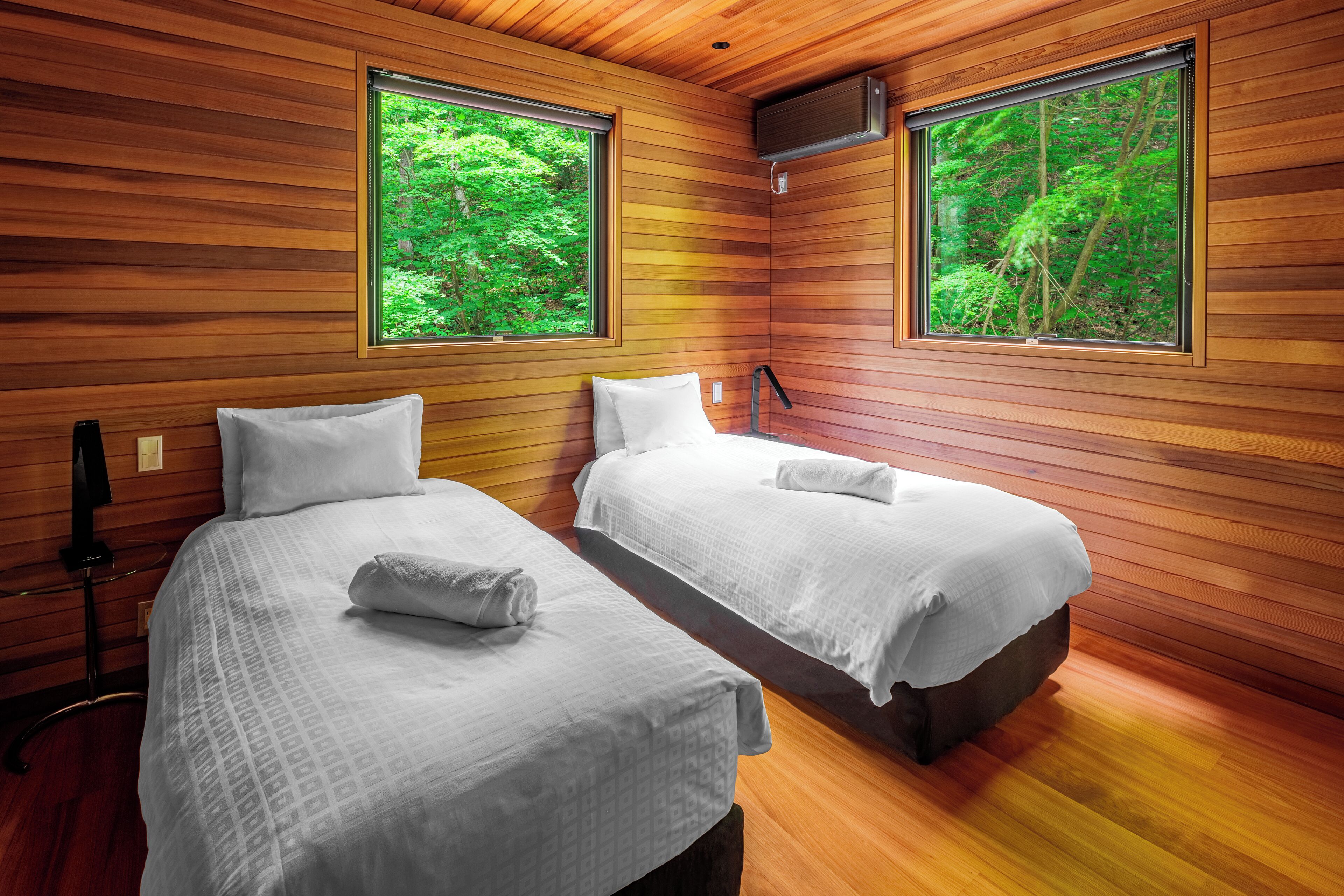 A room with two beds, wooden walls, and large windows with a forest view.