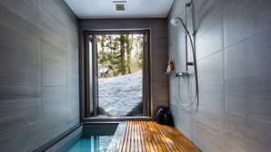A modern bathroom with a wooden floor, a glass-enclosed shower, and a view of a snowy landscape. - Phoenix Chalets (Hakuba)