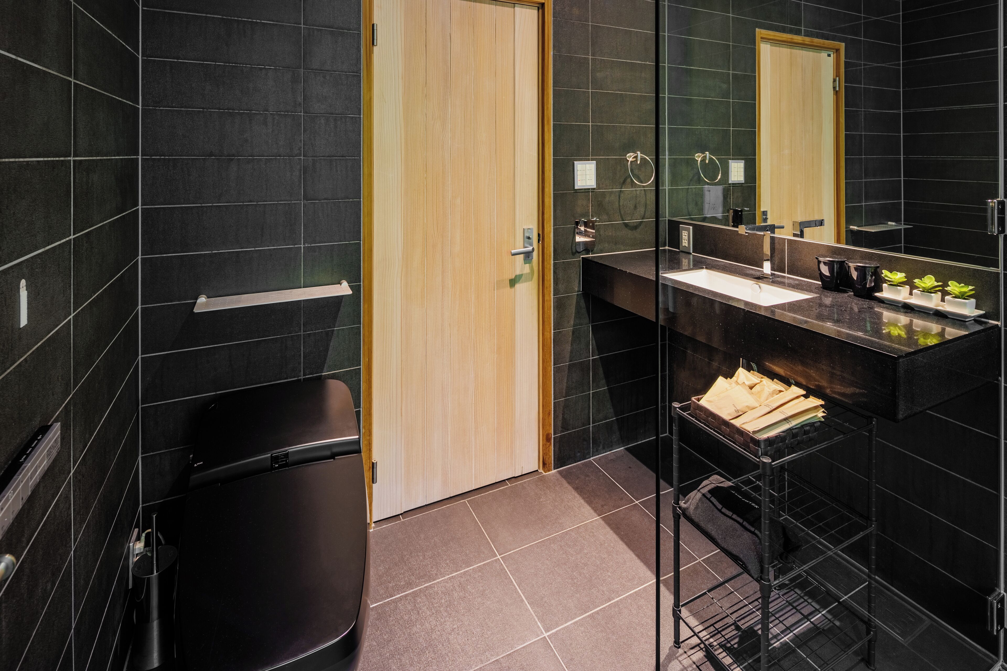 A modern bathroom with dark tiles, a wooden door, a black trash bin, a glass shelf with toiletries, and a large mirror.