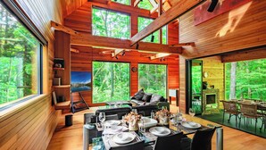 A wooden cabin with a dining area, a fireplace, and large windows overlooking a forest. - Phoenix Chalets (Hakuba)