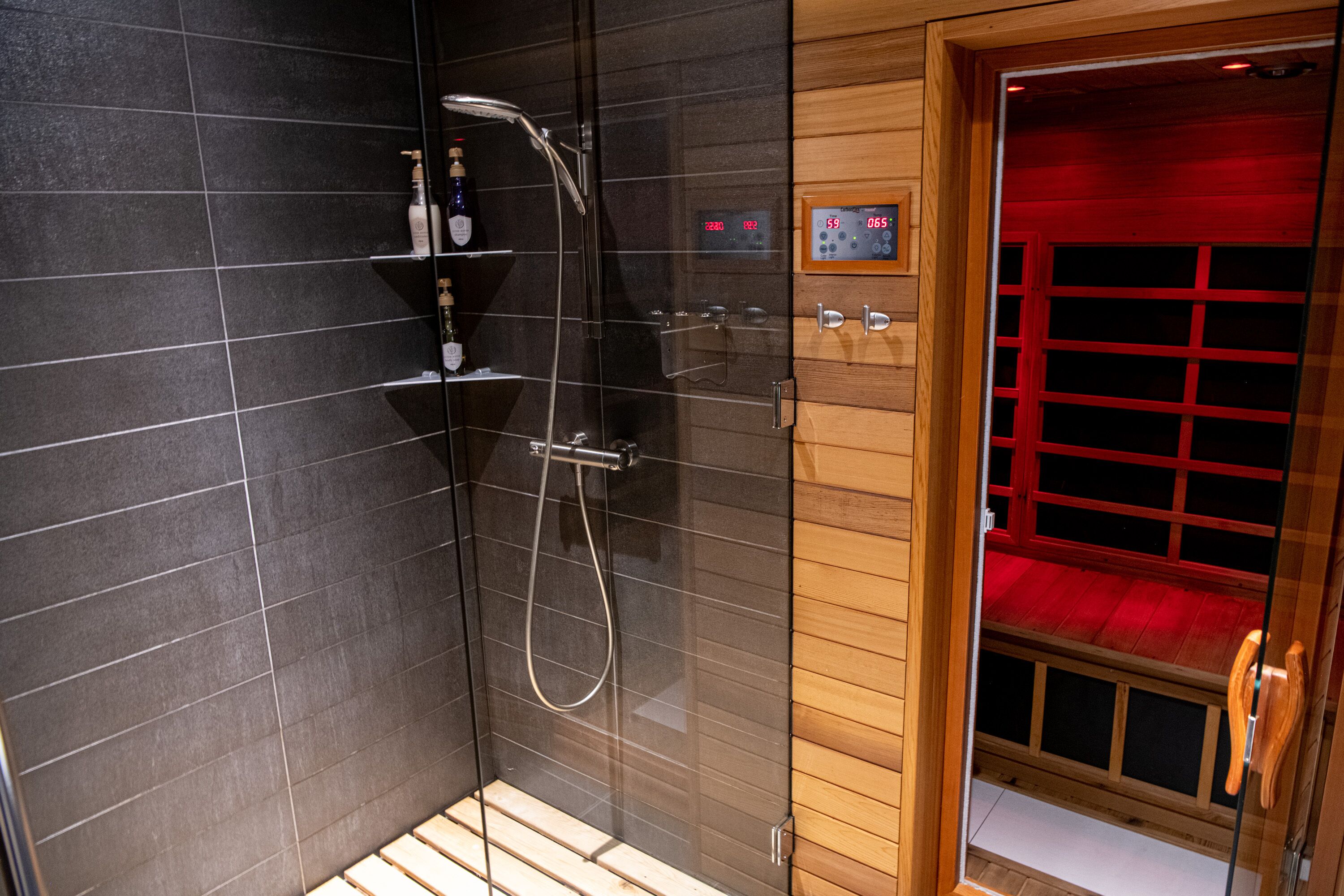 A modern shower area with a digital control panel, a glass-enclosed shower, and a wooden door leading to a red-lit room.