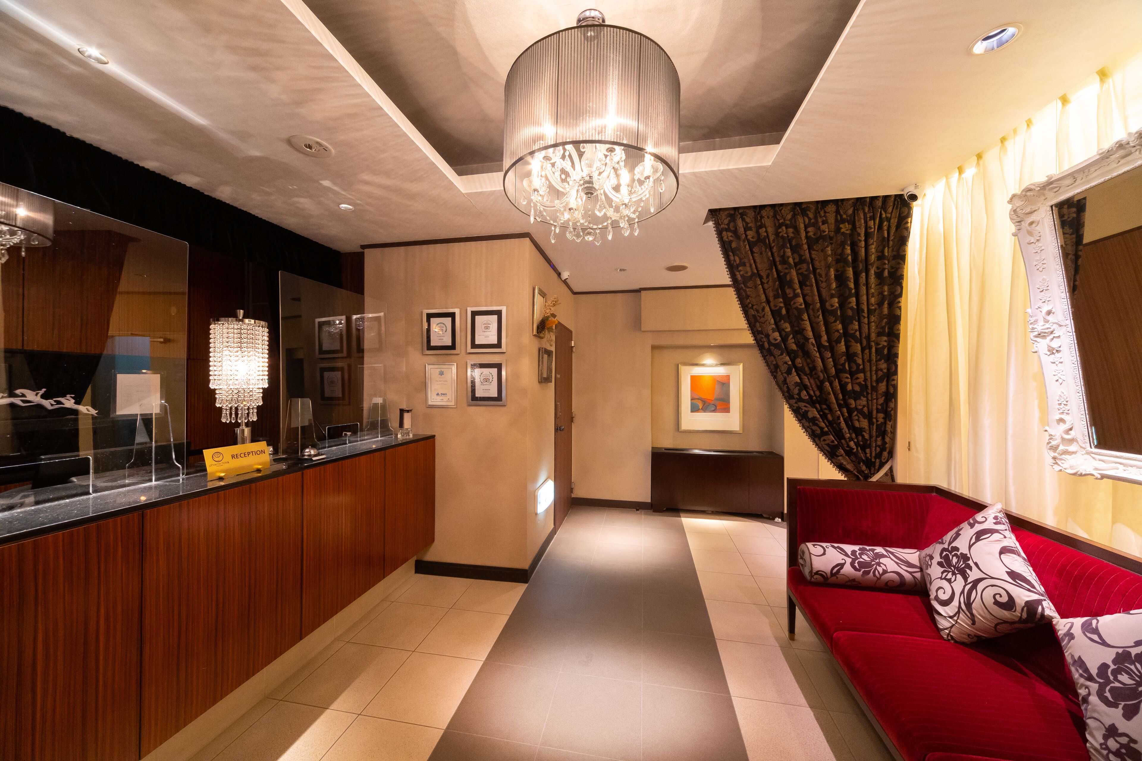 A hotel reception area with a reception desk, a chandelier, a red sofa, and a hallway leading to another room.