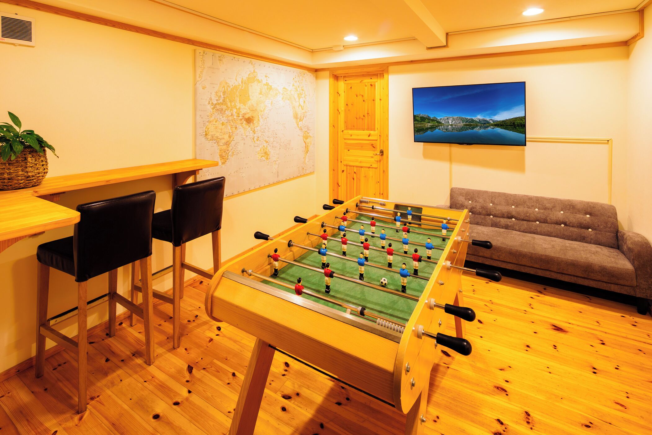 Una habitació amb taula de futbolí, dos tamborets, un sofà, televisió i mapa a la paret.