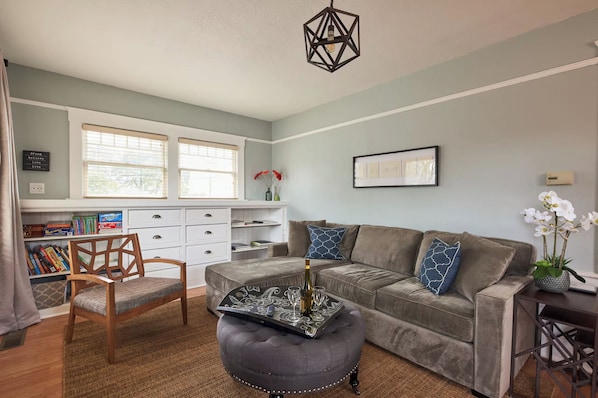 Una sala de estar con un sofá gris, una silla de madera, una mesa de centro con una bandeja decorativa, una lámpara colgante y una estantería con libros y objetos decorativos.