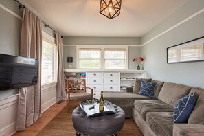 A living room with a sofa, ottoman, and a flat-screen TV. There is a dining area with a table and chairs, and a kitchen area with cabinets and a countertop.