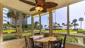 A dining area with a round table and chairs, large windows with a view of palm trees and buildings, and a ceiling fan. - Oceanfront/Beachfront -Sarasota Surf  Racquet - Y Building (Sarasota)