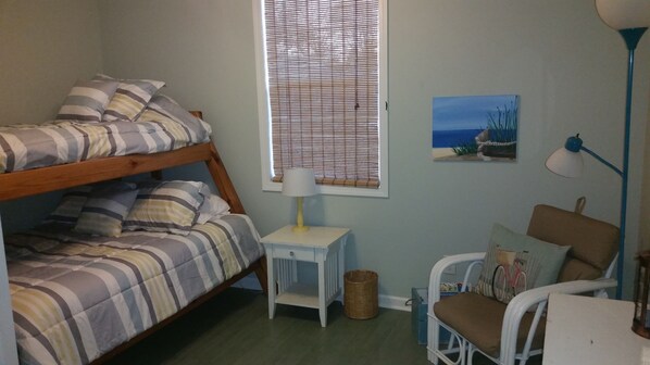 A bunk bed with striped bedding, a small white nightstand with a lamp, a wicker basket, a blue chair, and a wall painting of a beach scene. - The Lake House Cottage - Sleeps 5 (Fort Gratiot Township)