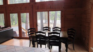 Una cabaña de madera con comedor, que incluye una mesa y sillas, ventanas amplias y vistas al bosque circundante.