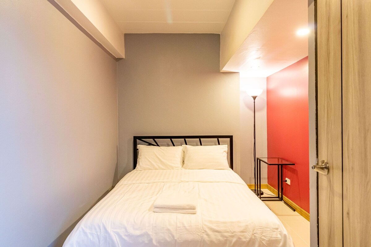 A small, neatly made bed with white linens in a room with a red accent wall and a wooden door.