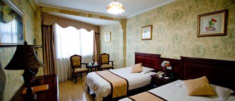 A hotel room with two beds, a chandelier, a desk with a lamp, a chair, a small table, and a window with curtains.