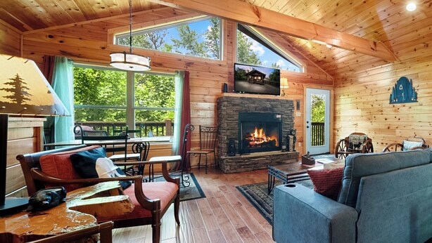 Interior acogedor de una cabaña con chimenea de piedra, paredes de madera y ventanas amplias que ofrecen vistas al exterior.