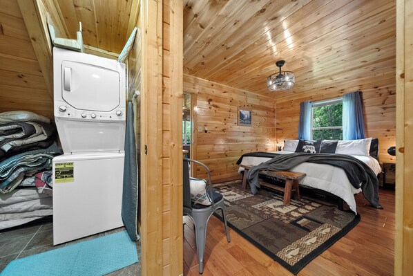 A wooden cabin interior with a bed, a chair, a small table, and a washing machine.