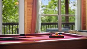 Una mesa de billar con bolas y tacos, una ventana con vistas a árboles y paredes de madera.