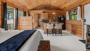 Un chalet confortable avec un intérieur en bois, un lit, un coin repas avec deux chaises, une cuisine équipée d’un réfrigérateur et une grande fenêtre.