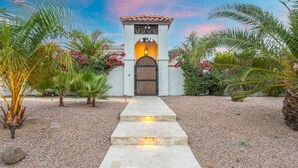 A pathway leading to a gated entrance with a light fixture above the door, surrounded by palm trees and flowering plants.