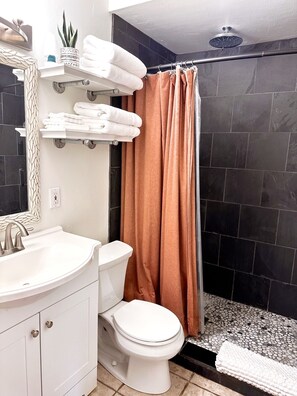 Un bagno con lavandino, water e tenda doccia bianca. Asciugamani su uno scaffale e una pianta in vaso.