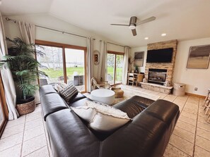 A spacious living room with a fireplace, leather sofas, a dining area, and a large window offering a view of the outdoors. - Door county cottage (Sturgeon Bay)