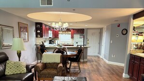 A living room with a sofa, ottoman, and a coffee table. There is a dining area with chairs and a table. The kitchen is visible in the background with cabinets and a refrigerator. - Cascading Creek / Gatehouse Condos / 2BR / 1st floor / no steps (Gatlinburg)