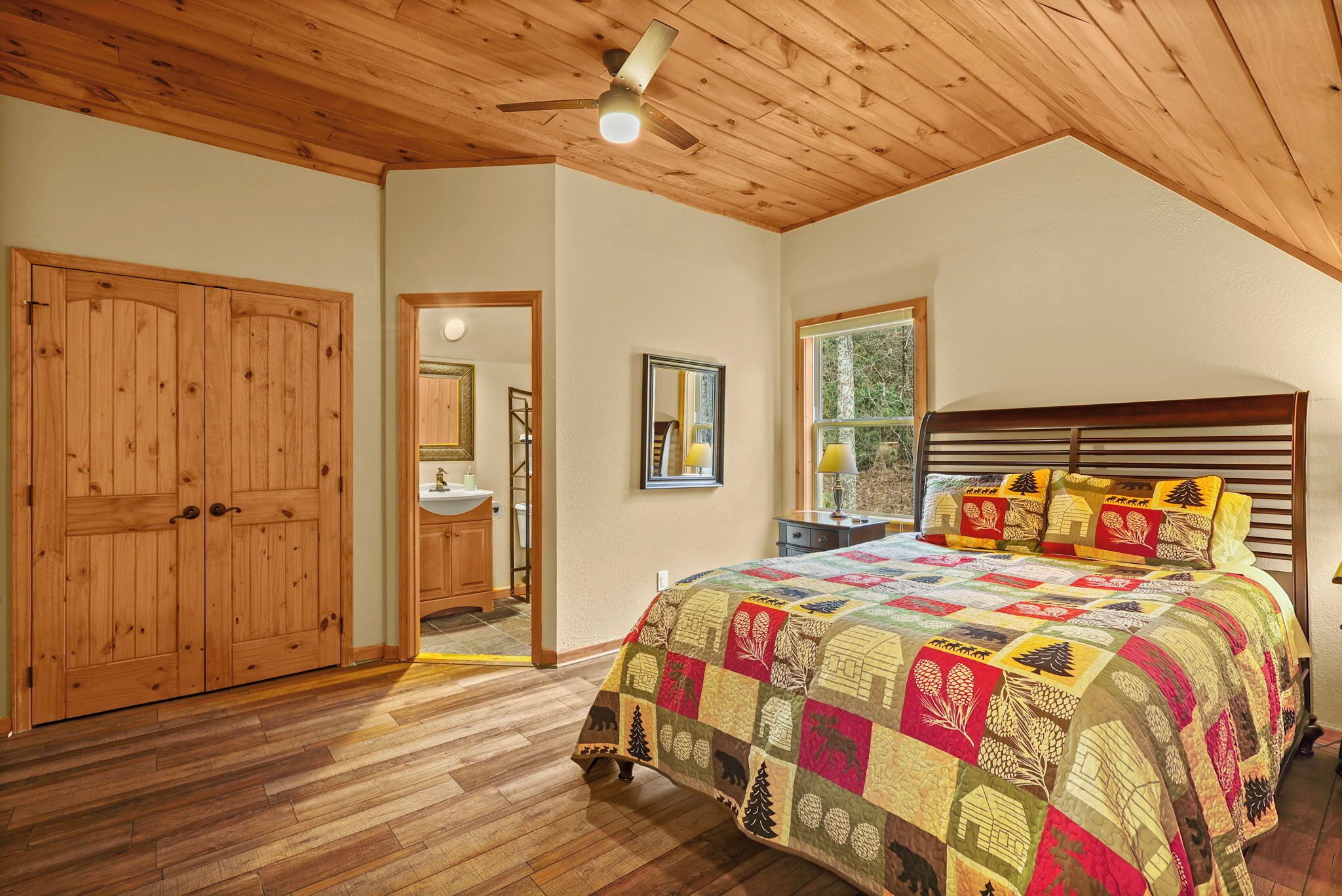 Une chambre avec un plafond en bois, un grand lit recouvert d’une couette aux couleurs vives, une tête de lit en bois et une fenêtre donnant sur les arbres.