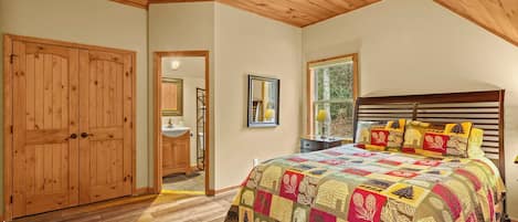 Une chambre avec un plafond en bois, un grand lit recouvert d’une couette aux couleurs vives, une tête de lit en bois et une fenêtre donnant sur les arbres.