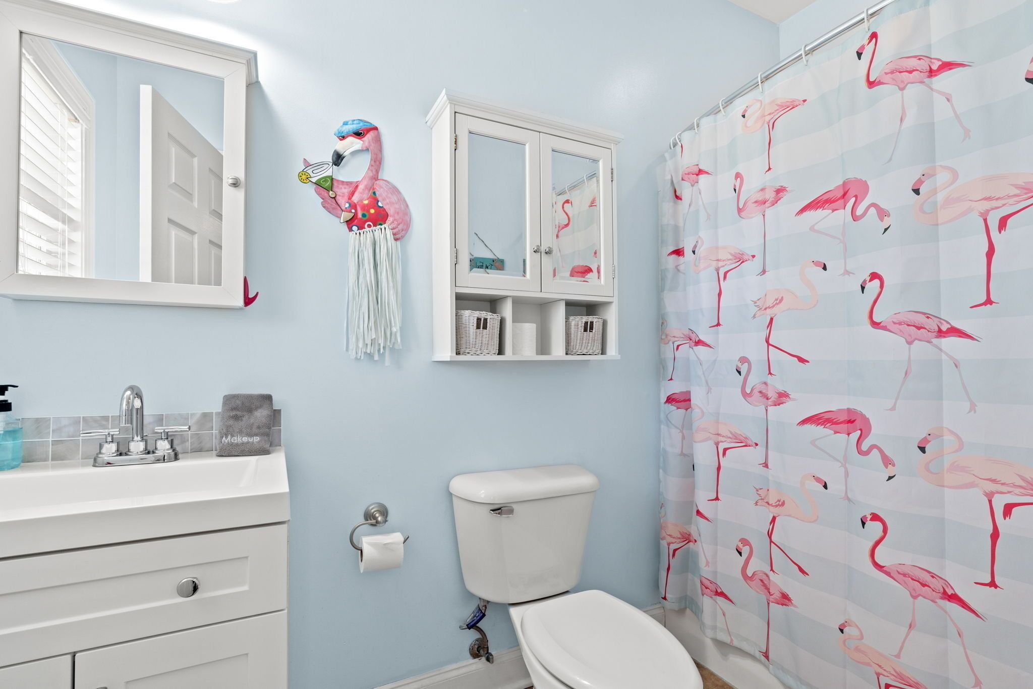 Une salle de bain avec des toilettes, un lavabo et un miroir. On y trouve un meuble avec des paniers et un flamant rose décoratif accroché au mur.