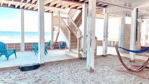 Ein Strandhaus mit Holzterrasse, Sitzgelegenheiten im Freien und einer Treppe, die zum oberen Stockwerk führt.