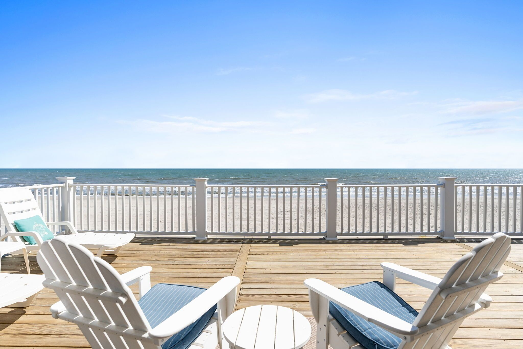 Une terrasse en bois, agrémentée de chaises Adirondack blanches et d’une petite table ronde, qui offre une vue sur une plage de sable et l’océan.