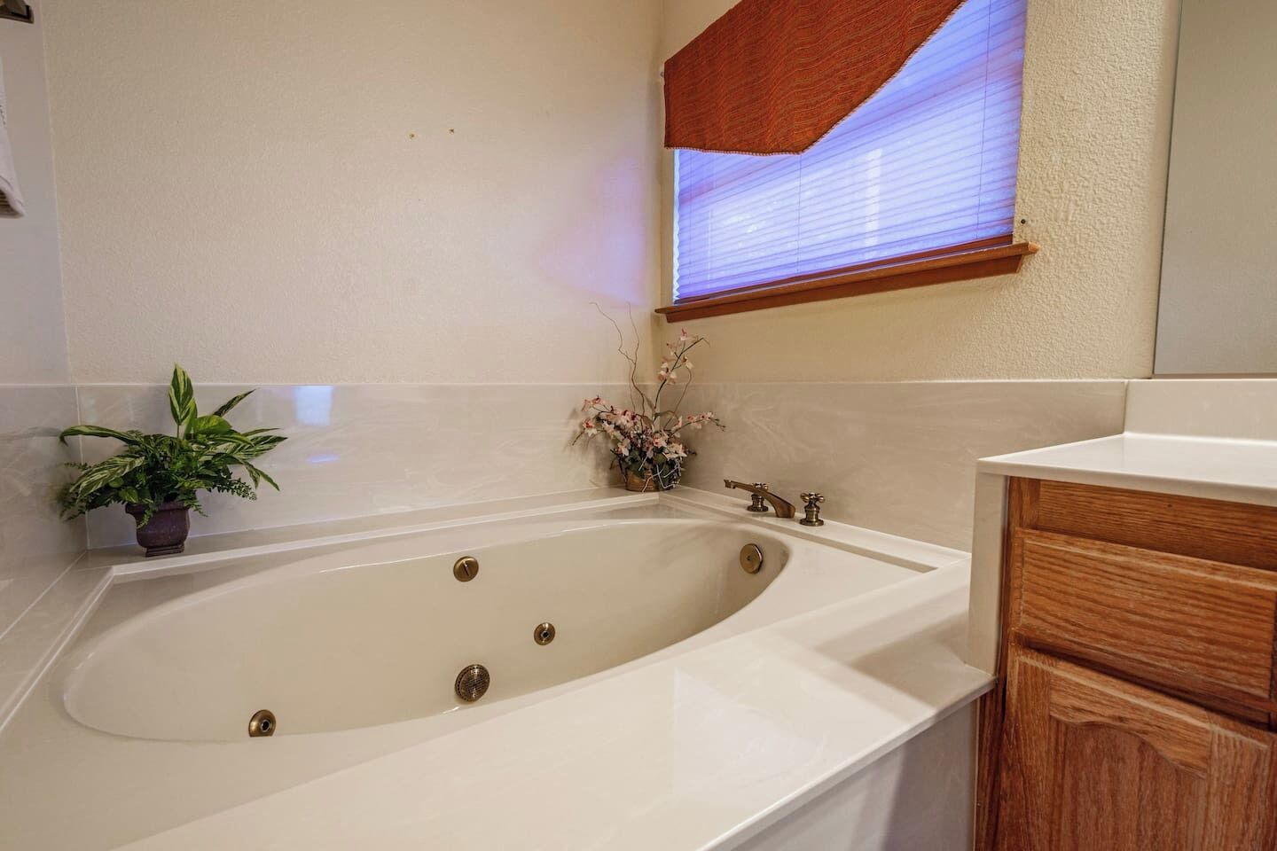 A bathroom with a freestanding bathtub, a potted plant, a vase with flowers, and a window with blinds.