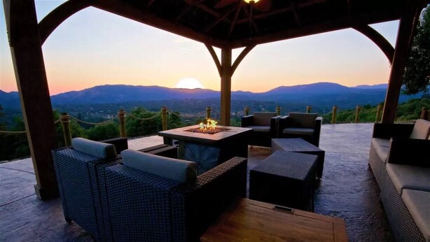 An outdoor seating area with wicker furniture and a wooden table, overlooking a mountainous landscape at sunset.