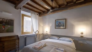 A bedroom with a wooden ceiling, a bed with a striped bedspread, a wooden dresser, a window with curtains, and two wall-mounted lamps. - Cosy Medieval Townhouse, between lake, sea, thermal baths, Rome, Viterbo, Umbria (Anguillara Sabazia)