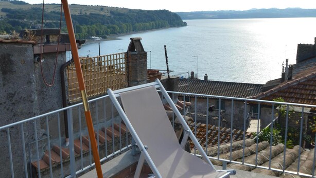 A balcony with a white sun lounger and a large umbrella, overlooking a body of water and hills. - Cosy Medieval Townhouse, between lake, sea, thermal baths, Rome, Viterbo, Umbria (Anguillara Sabazia)