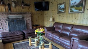 A cozy living room with a brown leather sofa, a glass coffee table with flowers, a fireplace, and wooden walls. - CASCADE RETREAT MOUNTAIN VIEWS, GREAT LOCATION NEAR COLORADO SPRINGS ATTRACTIONS (Cascade-Chipita Park)