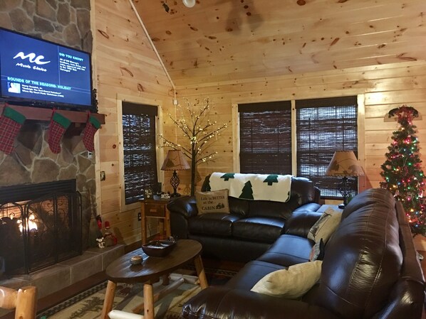 Interior acogedor de una cabaña con chimenea de piedra, sofá de cuero, adornos navideños y un televisor reproduciendo música festiva.