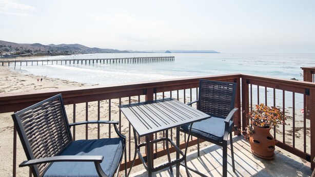 Une terrasse au bord de la plage, avec deux chaises en métal et une table, d’où l’on peut admirer une plage de sable et une jetée qui s’avance dans l’océan.