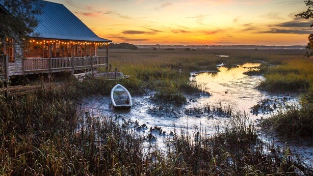 Une cabane rustique dotée d’une véranda et illuminée de guirlandes lumineuses, située près d’une zone marécageuse, avec un petit bateau à proximité.