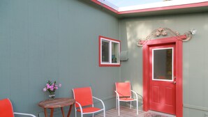 Eine Veranda mit zwei orangefarbenen Stühlen, einem kleinen runden Tisch und einer roten Tür mit einem dekorativen Bogen.