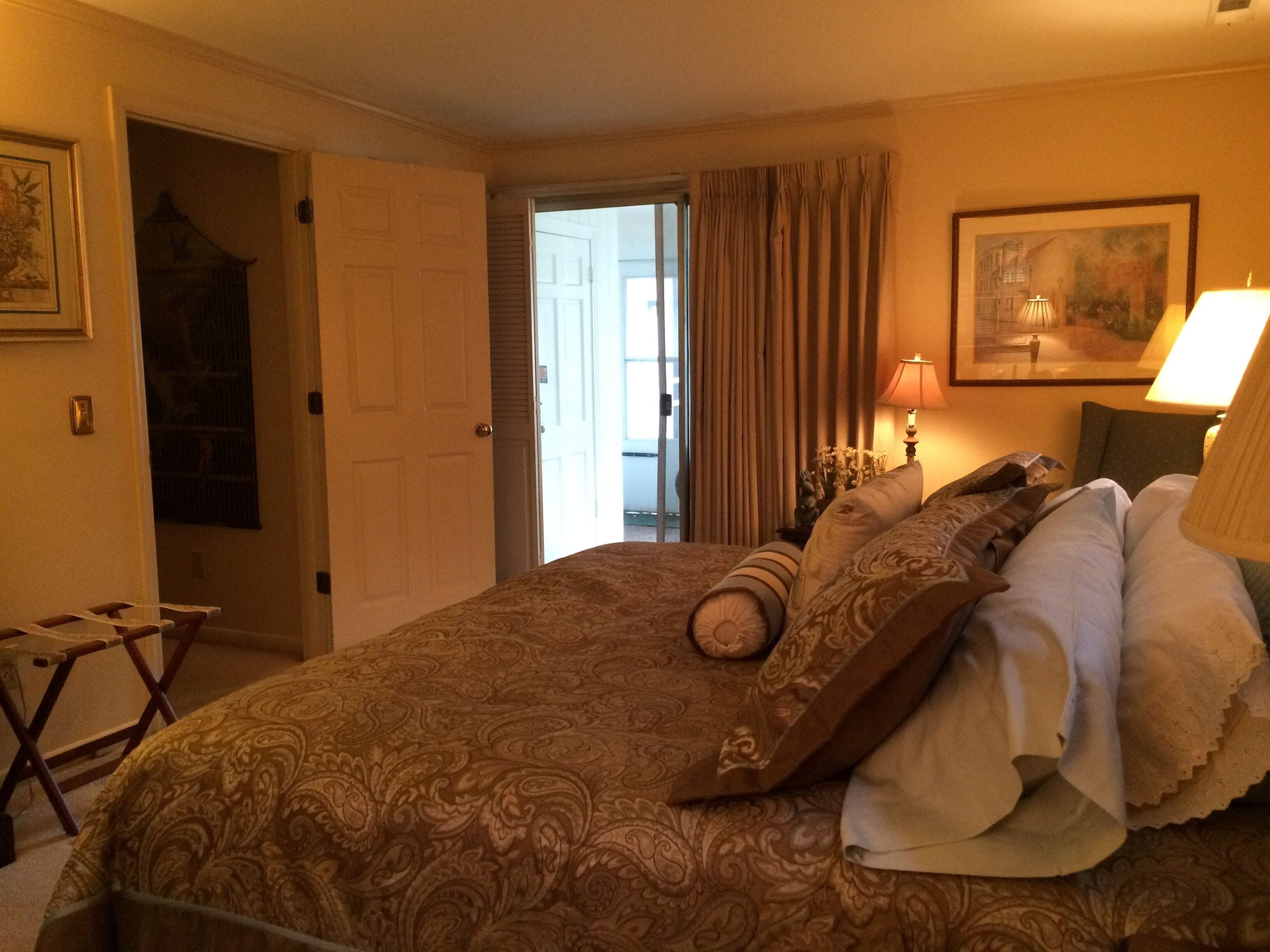 A hotel room with a large bed, a door leading to a balcony, a framed picture on the wall, and a nightstand with a lamp.