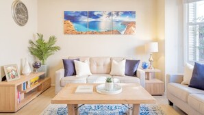 Ein Wohnzimmer mit einem beigen Sofa, einem Couchtisch, einem Beistelltisch mit Lampe, einer Wanduhr und einem großen Landschaftsbild.