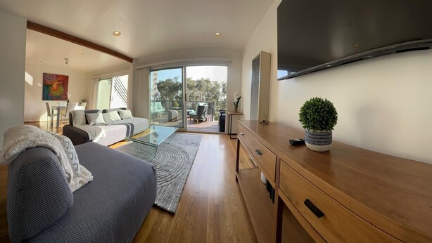 A modern living room with a sofa, coffee table, and a wooden desk. There is a large window and a balcony visible. - Beachy Private Get-A-Way!! Large Deck facing Ocean! (Playa Del Rey)