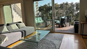 A modern living room with a glass coffee table, grey sofa, and a view of an outdoor deck with furniture. - Beachy Private Get-A-Way!! Large Deck facing Ocean! (Playa Del Rey)