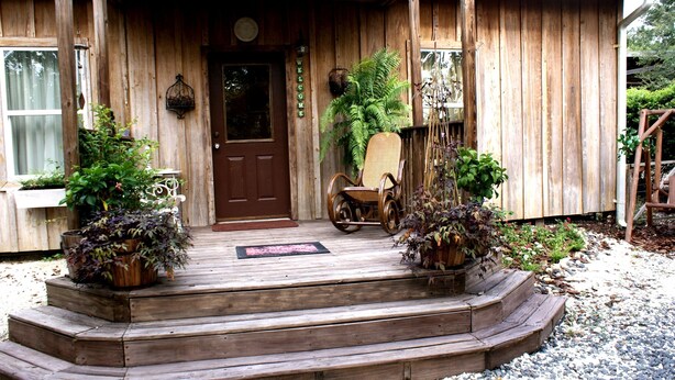 A wooden porch with a brown door, a wicker chair, and potted plants. - The Shebeen A small Irish cottage,come experience the enchantment and trnquility (Brooksville)