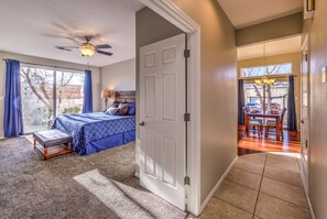 A bedroom with a bed, a bench, a ceiling fan, and a view of a dining area through an open door.