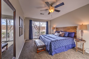 A bedroom with a large bed, a ceiling fan, a bedside table with a lamp, and a view through a sliding glass door.