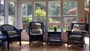 Ein sonnendurchfluteter Raum mit drei Korbstühlen und einem kleinen Tisch, große Fenster mit Blick auf einen Garten und ein Gebäude.