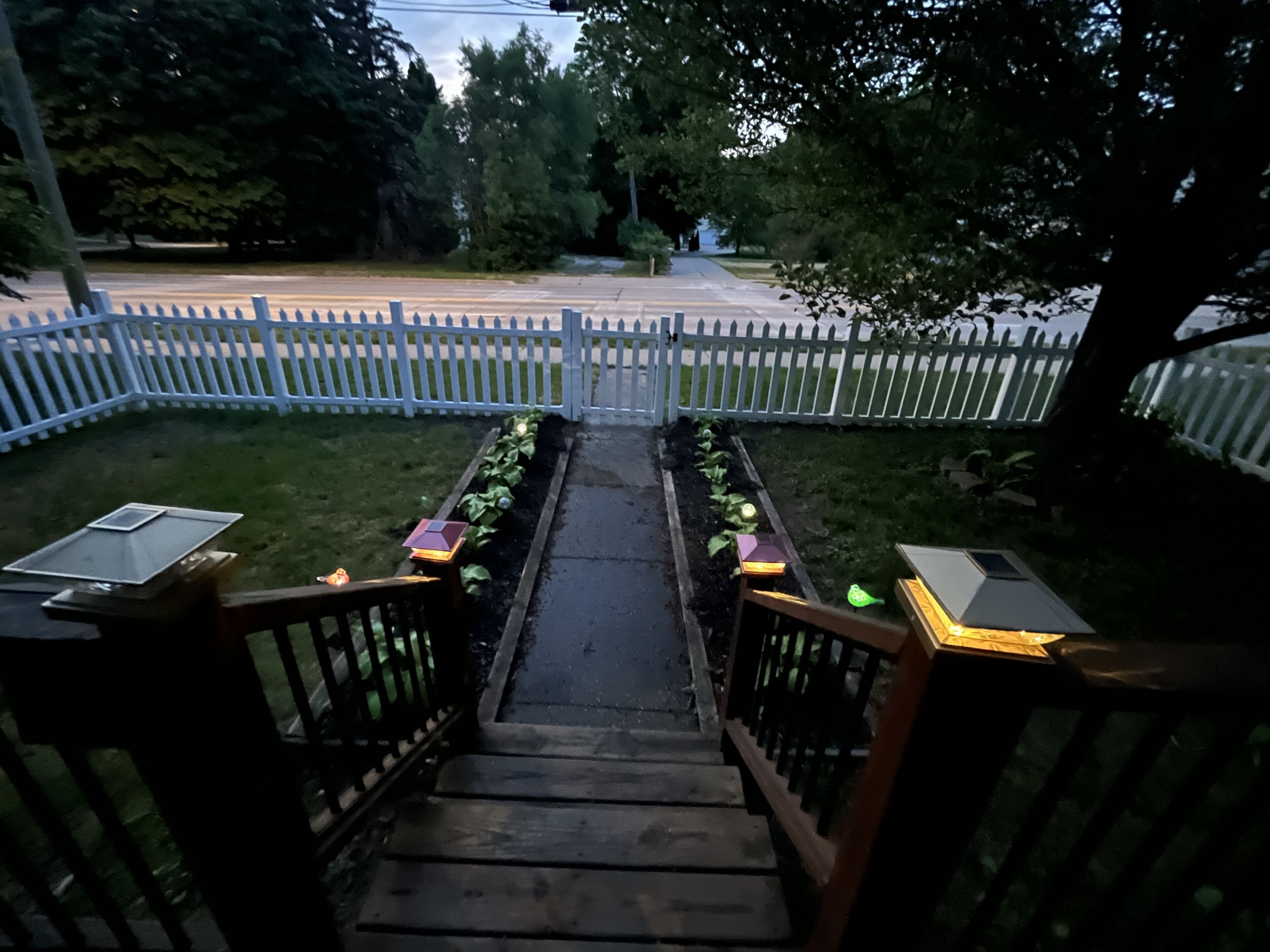 A wooden staircase with a white picket fence on both sides leading to a garden area with plants and solar-powered lights.
