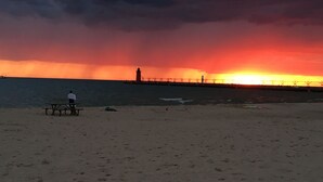 Ein Strand mit einer Person, die auf einer Bank sitzt, einem Pier im Hintergrund und einem Sonnenuntergang über dem Wasser.
