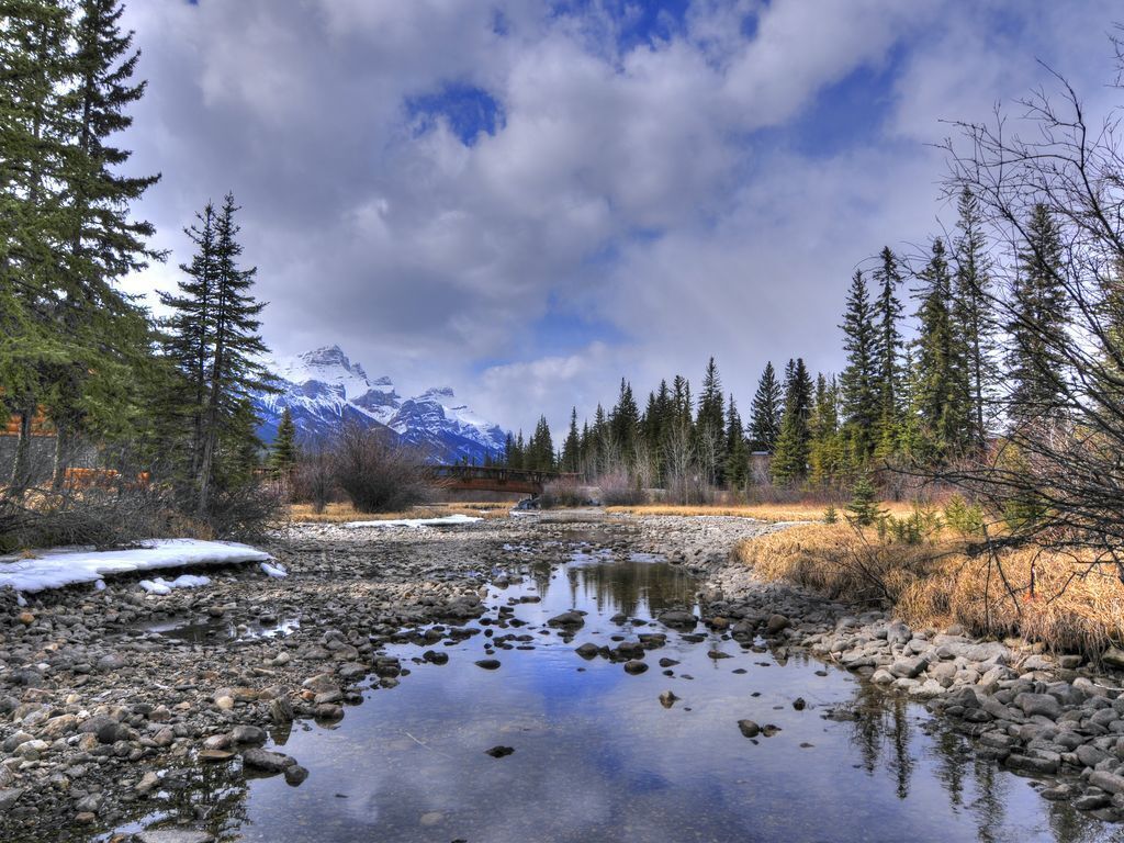 A river with rocks and trees, a mountain range in the background, and a cloudy sky.